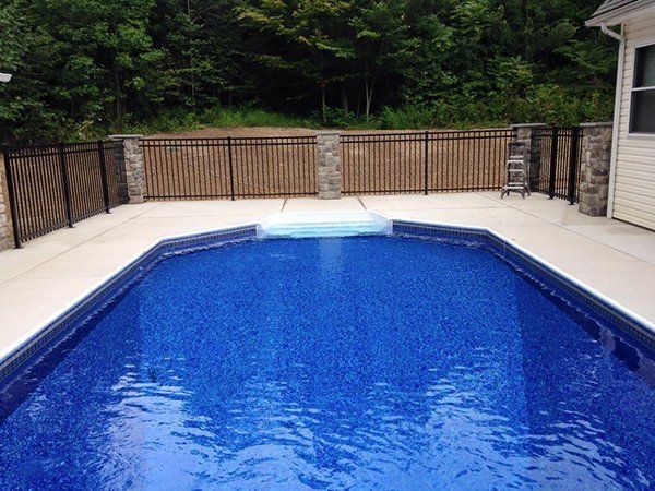 Outdoor swimming pool with bright blue water, tan deck, and black fence backed by trees