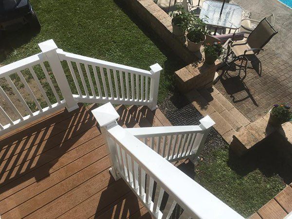View from a deck with white railings, wooden steps, and a patio with chairs and brick pavers below