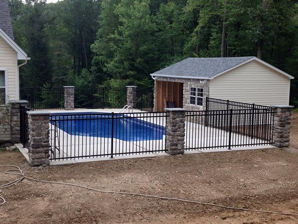 Backyard pool surrounded by black fence and stone pillars, with a small shed and wooded background.