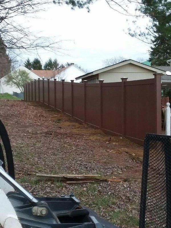 Brown privacy fence along a sloped yard beside a driveway and beige house under cloudy sky
