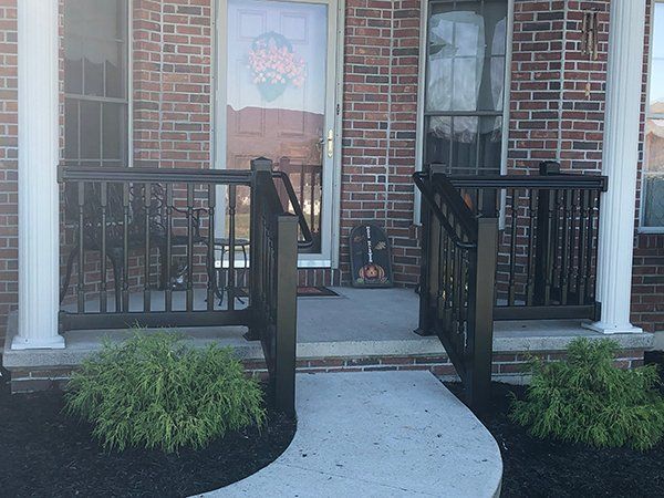 Front porch entrance with black railings, brick facade, white columns, and potted shrubs.