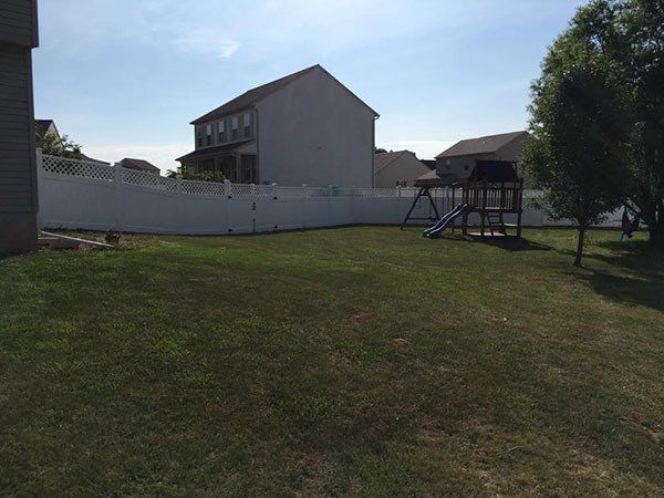 Backyard with a grassy lawn, white fence, house, and a playset under a partly sunny sky