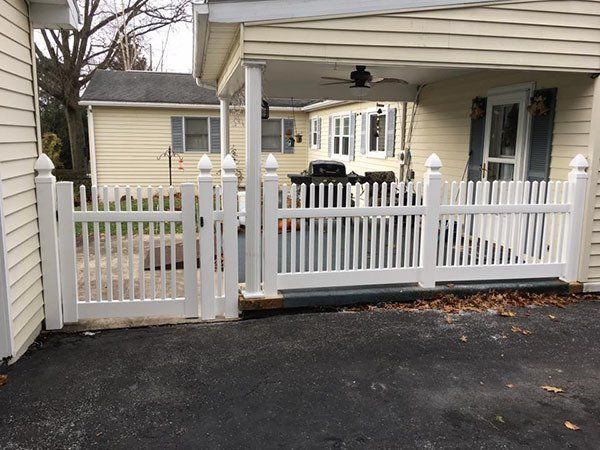 White picket gate and fence beside a beige house with a covered porch and black driveway.