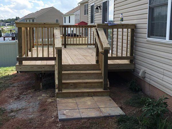 Wooden backyard porch with central steps and railings beside a house, leading to a paved landing.
