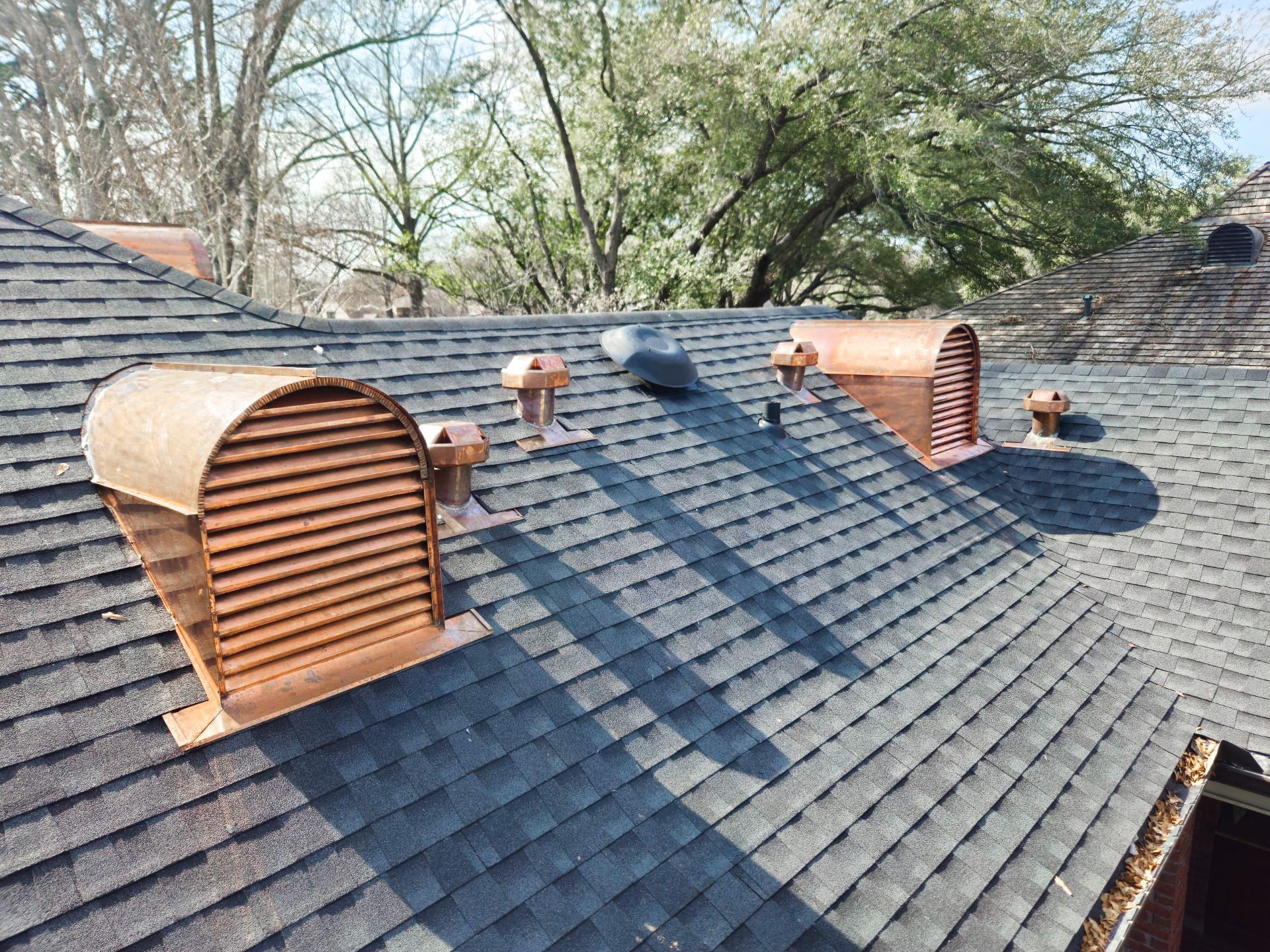 A dark shingled roof with copper vents and exhaust pipes, set against a backdrop of trees.
