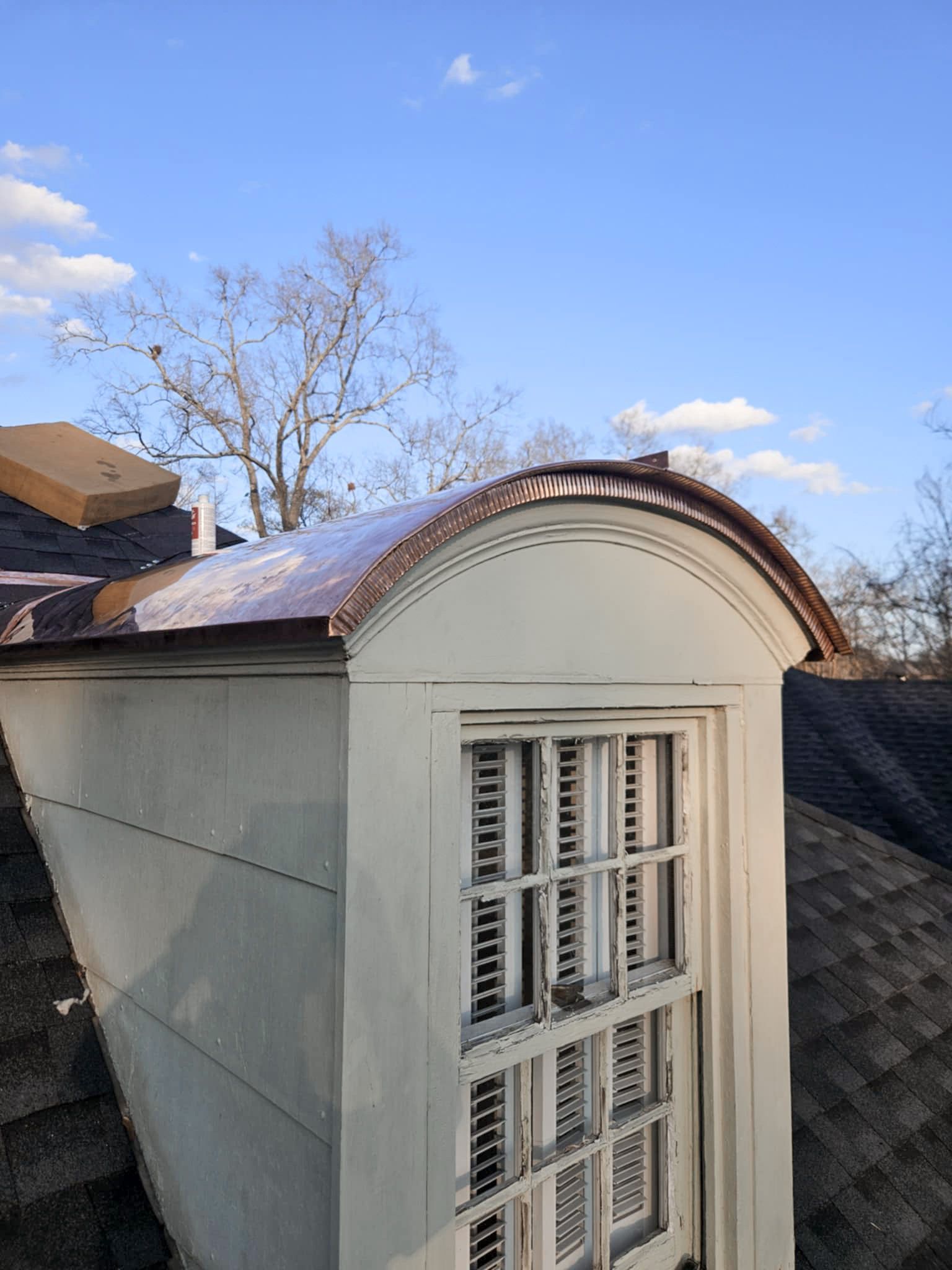 Dormer window on a house with a curved copper roof trim. The window has shutters and a blue sky background.