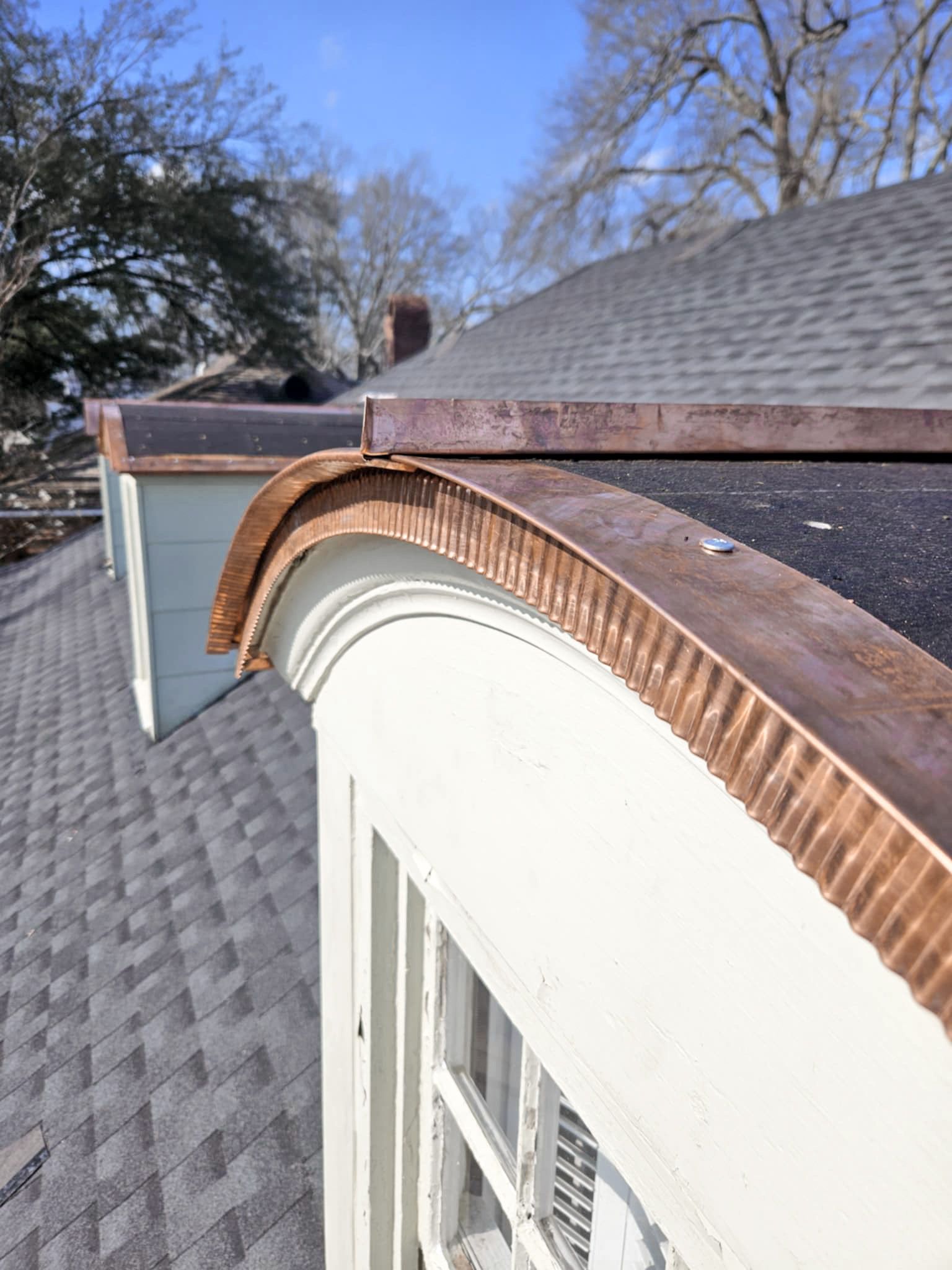 Copper roof trim on a white building with a curved architectural detail, set against a gray shingled roof and a blue sky.