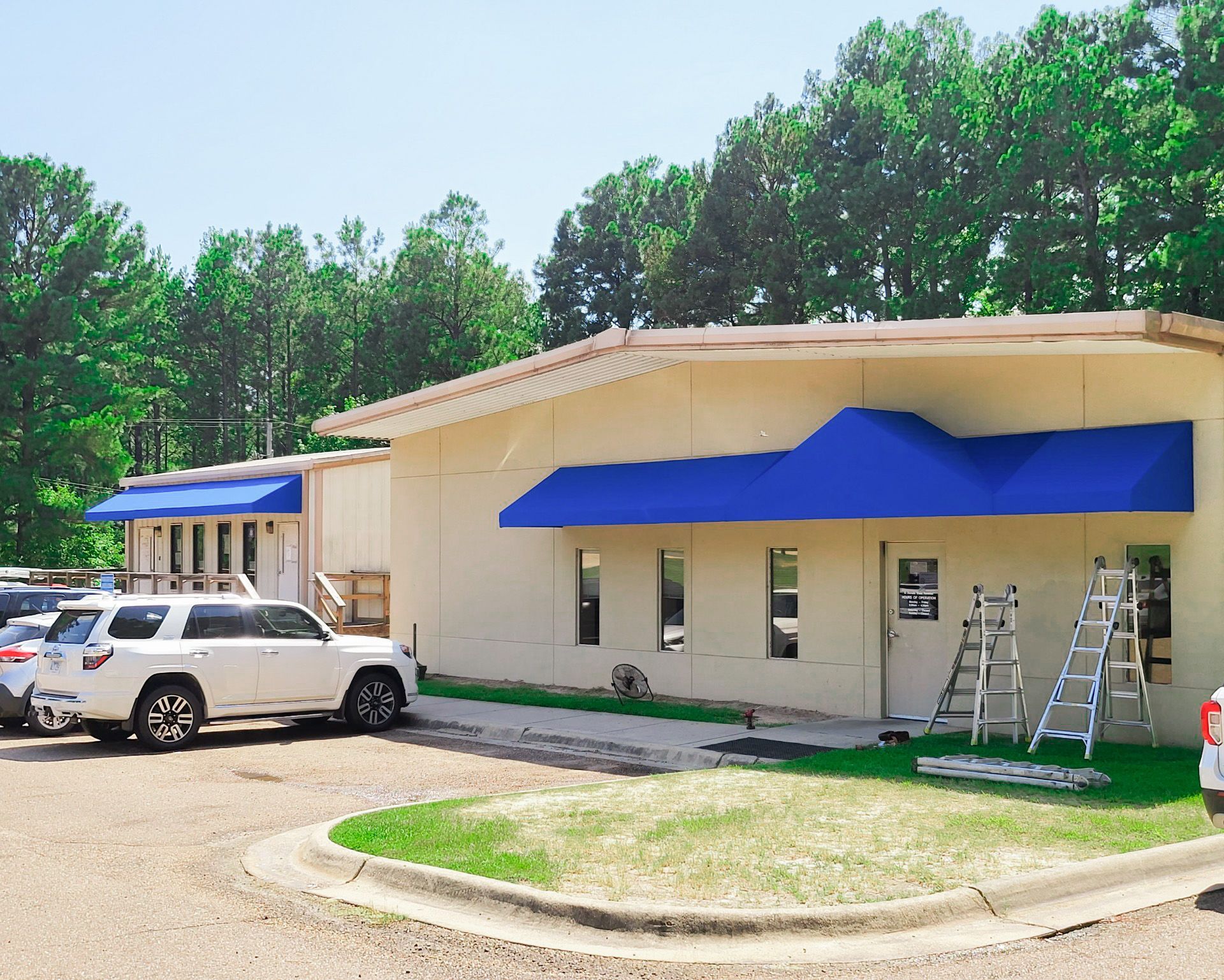 Exterior of a light-colored building with blue awnings; white SUV parked in front. Two ladders are leaned against the building.