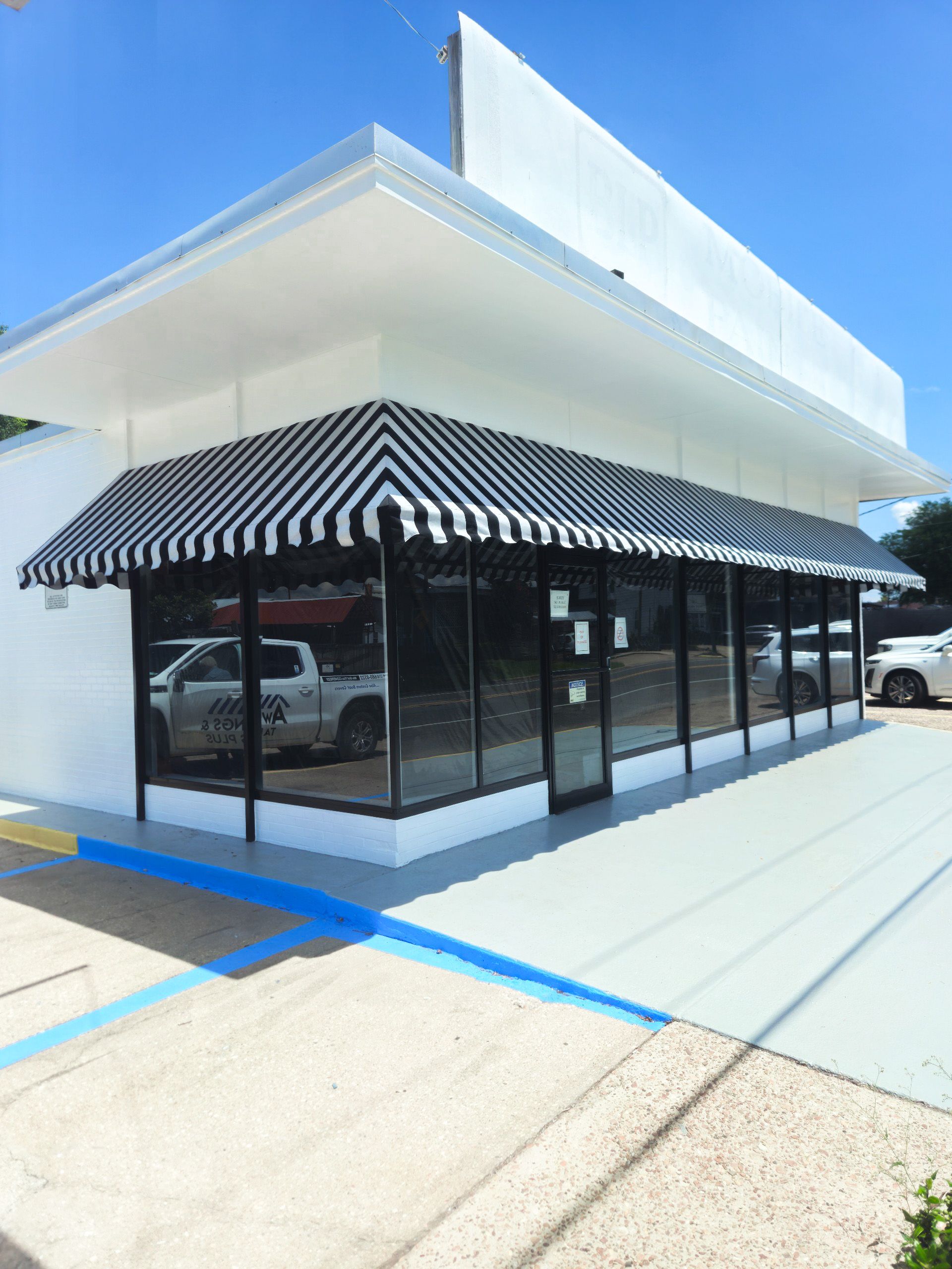 White building with black and white awning, large windows, parking spot, and a blue curb.
