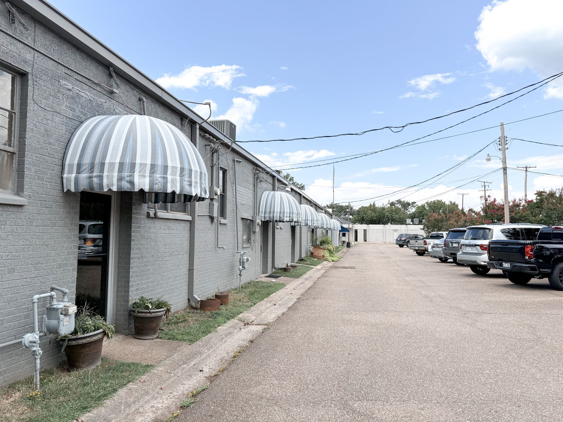 Grey building with striped awnings, doors, and a parking lot on a sunny day.