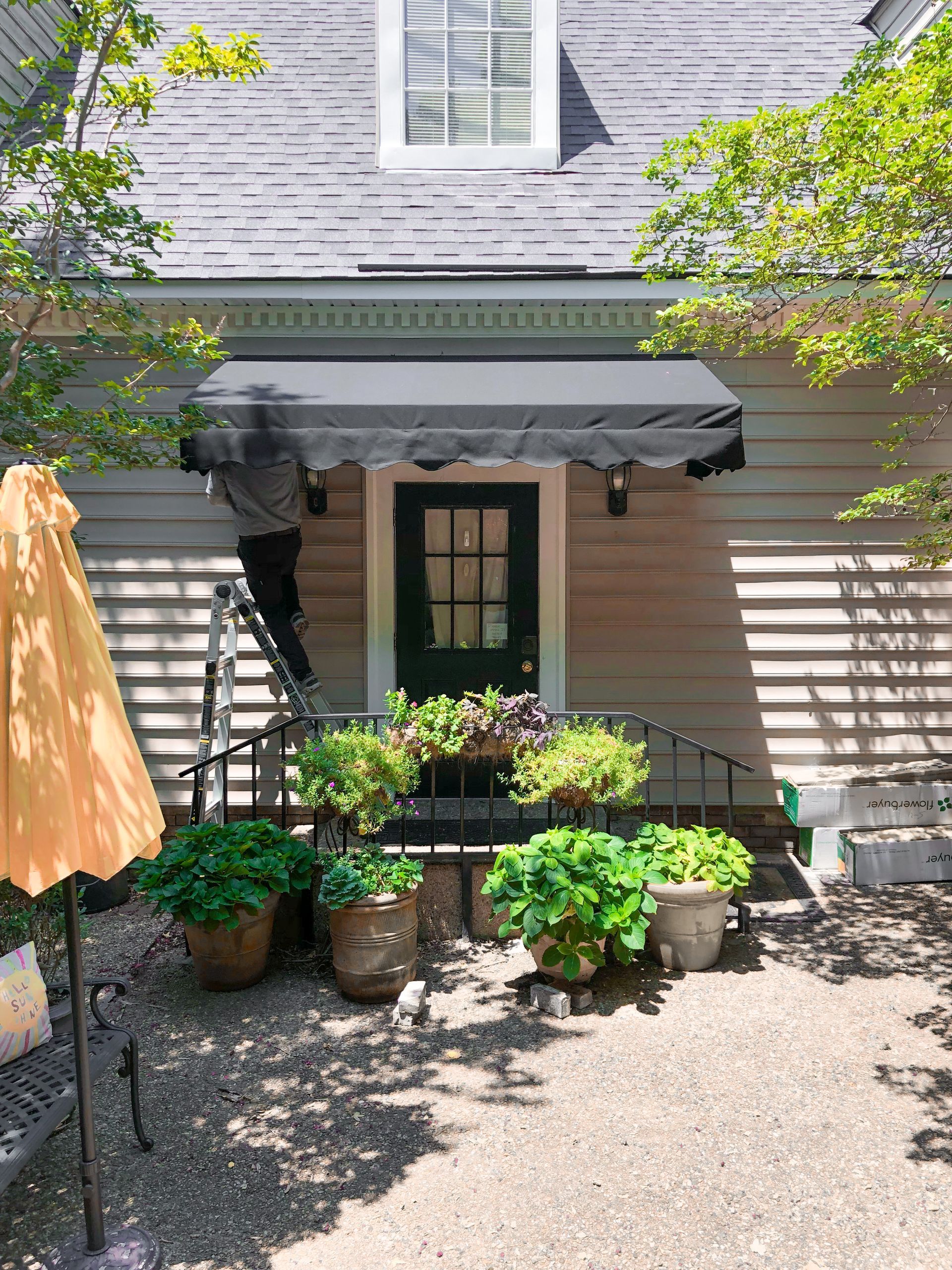 Person on a ladder adjusting a black awning over a green door, flanked by potted plants.