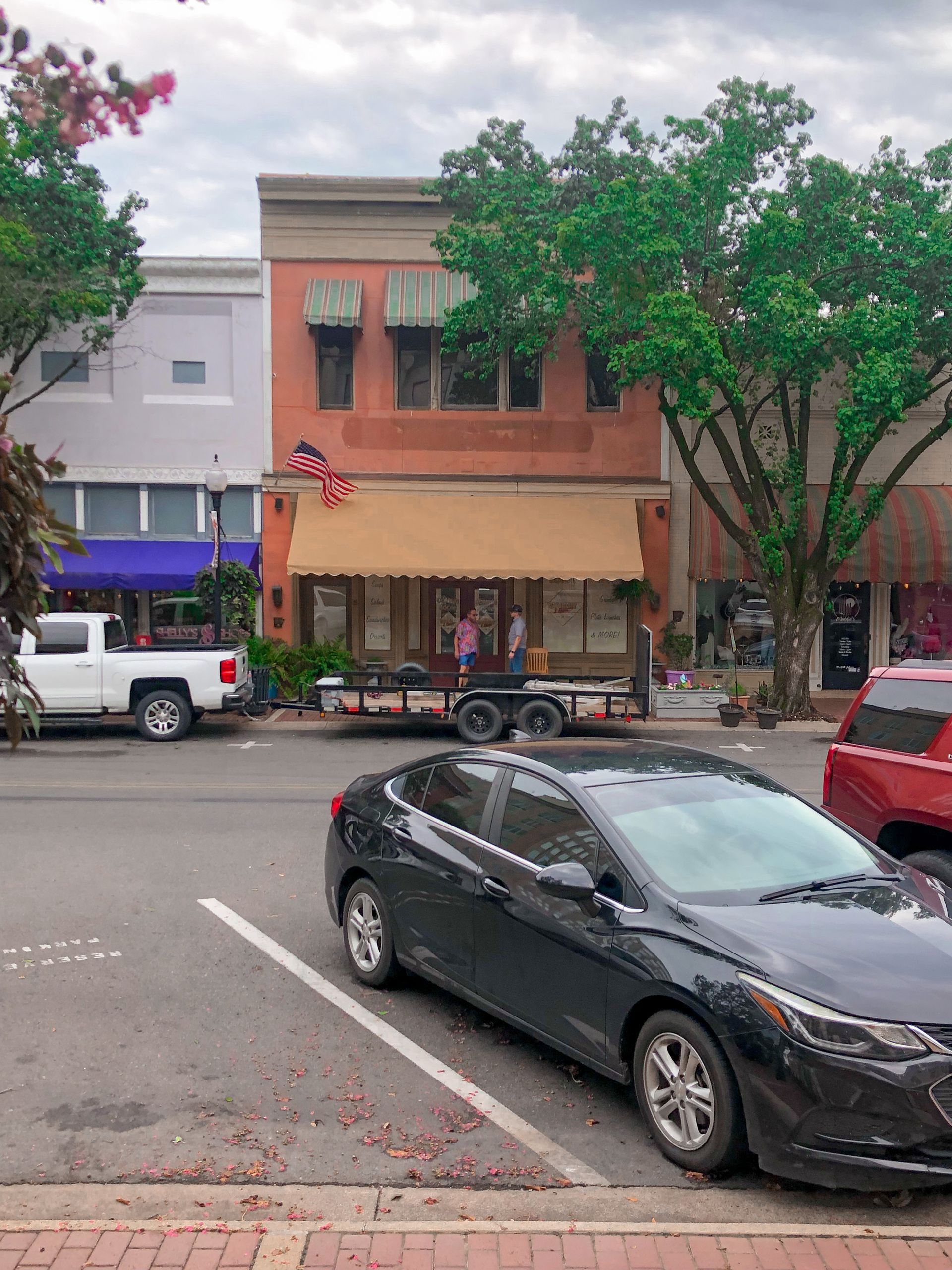 Street scene: red brick building with tan awning, American flag. Black car parked, white truck, trees.