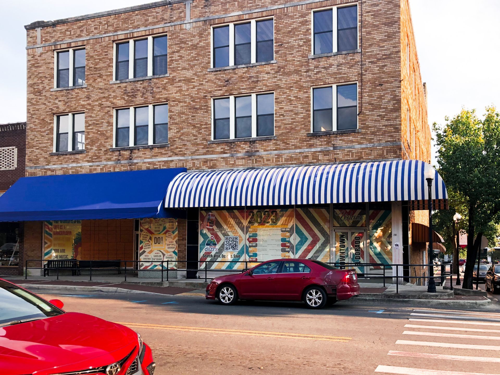 Three-story brick building with awnings, colorful mural, and red cars parked on the street.