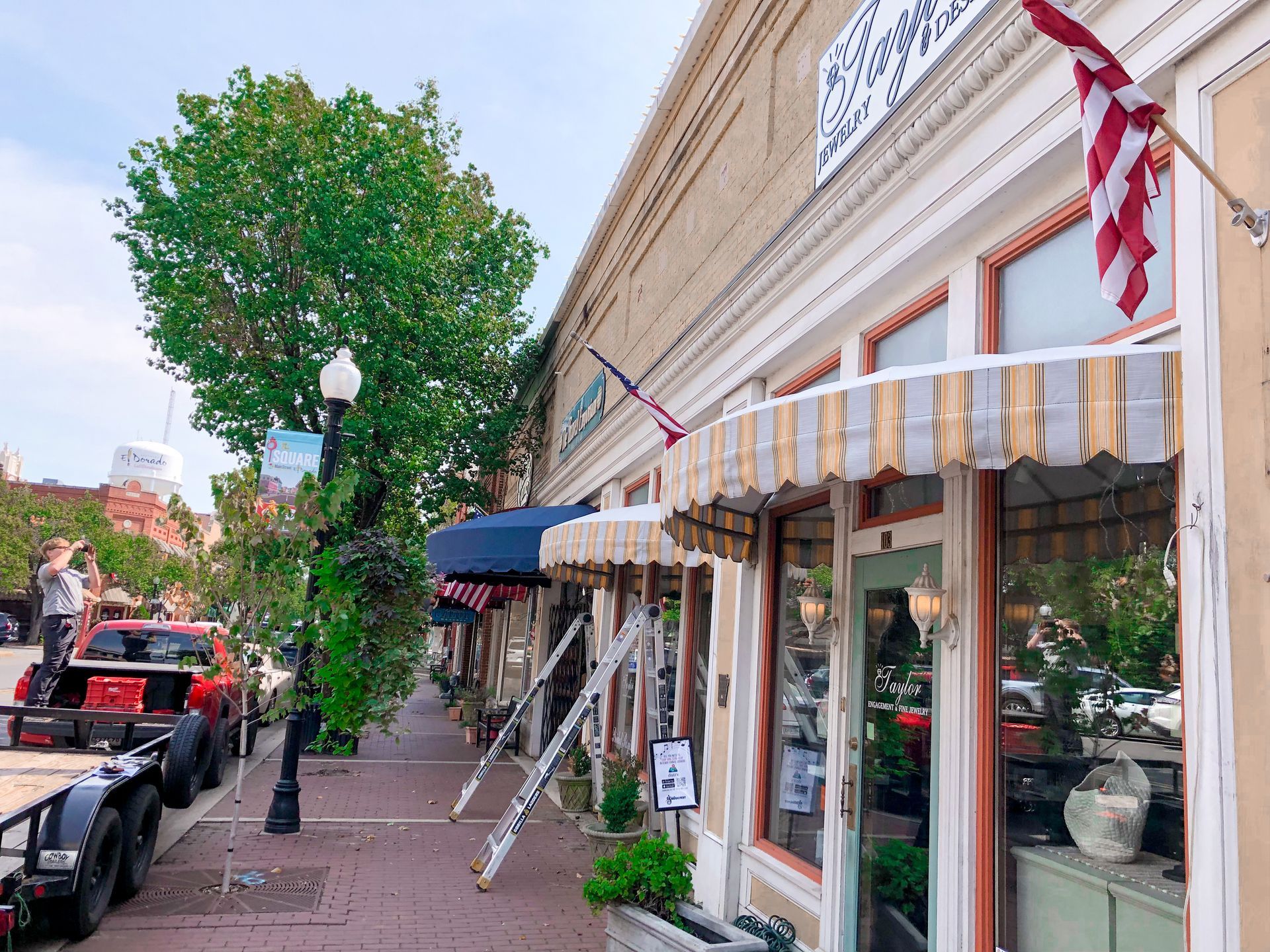 A downtown street with brick buildings and awnings; work being done in front of a shop, flags fly.
