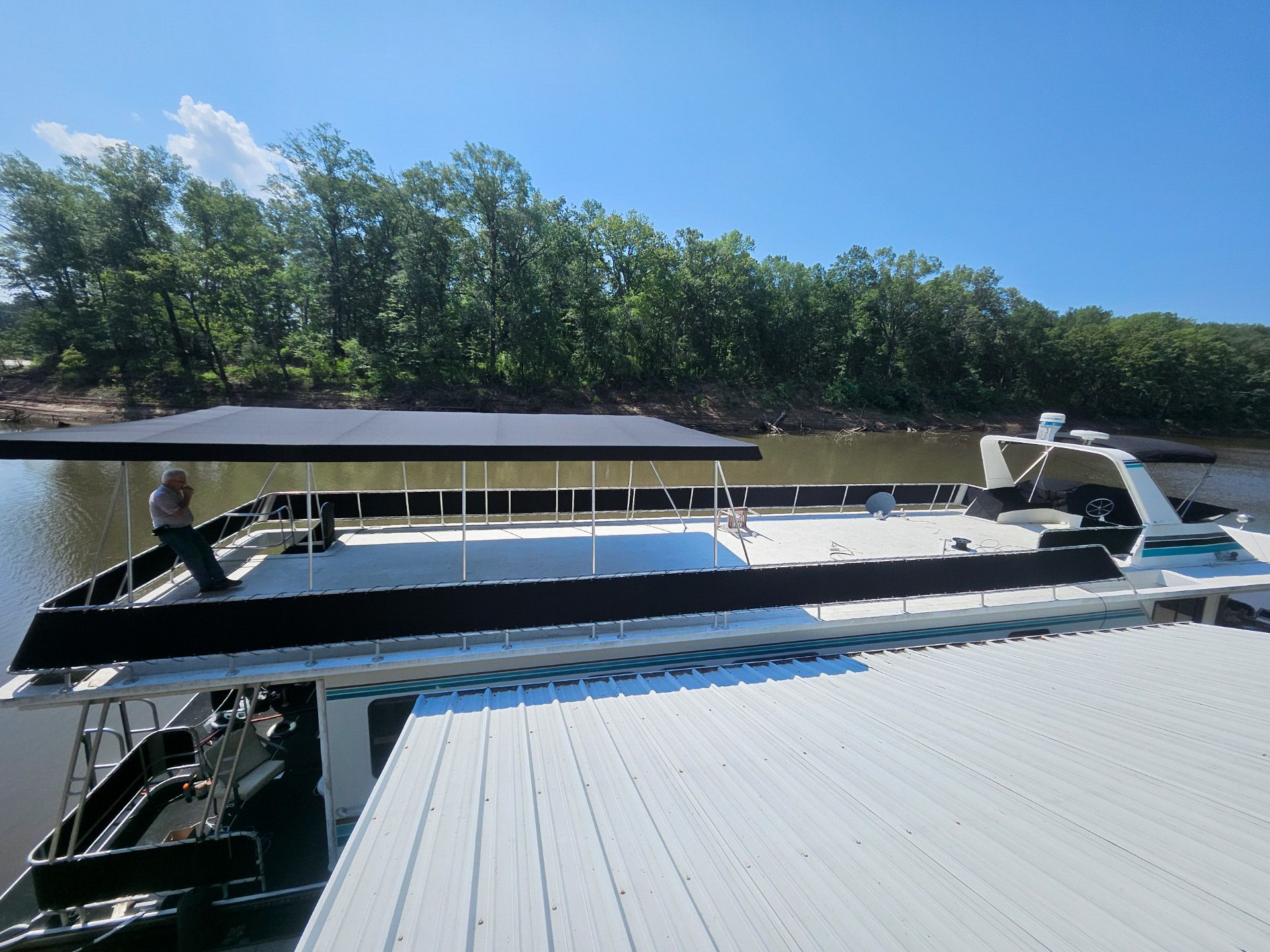 A pontoon boat on a river, with a person and trees in the background under a blue sky.
