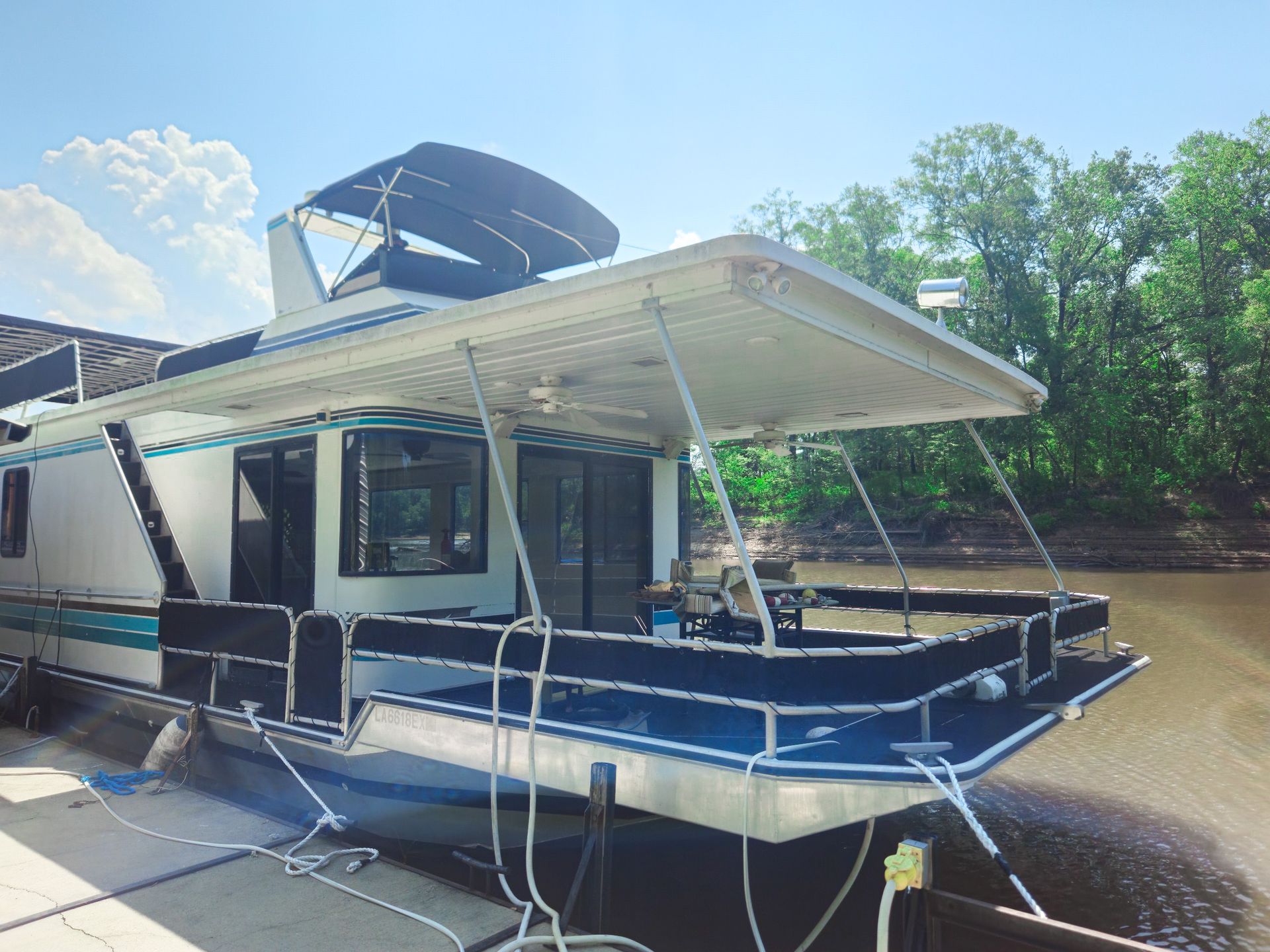 Houseboat docked on a river, white with blue trim, awnings, sunny day.