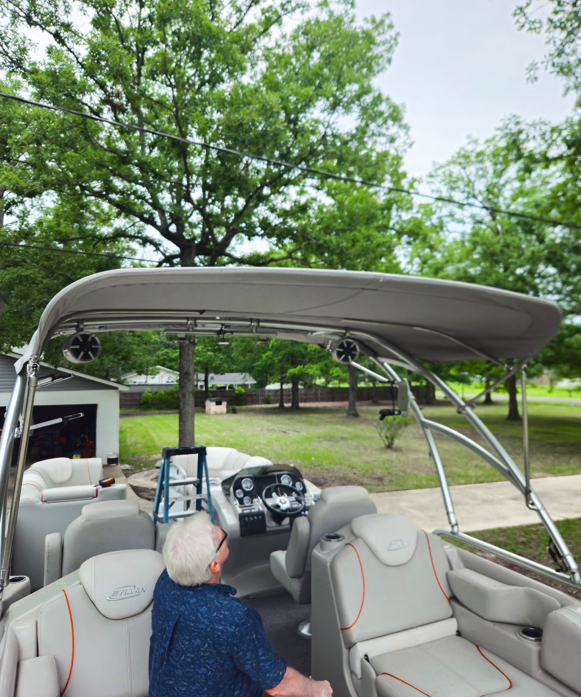 Senior sits in a pontoon boat under a canopy on a cloudy day; trees in background.