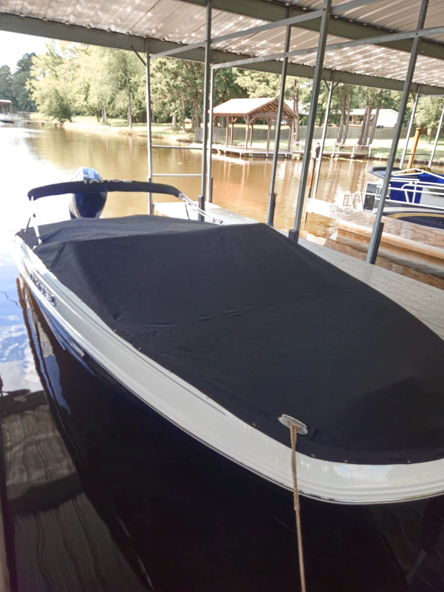 Black boat with cover docked at a pier, under a covered structure. Water, trees in background.