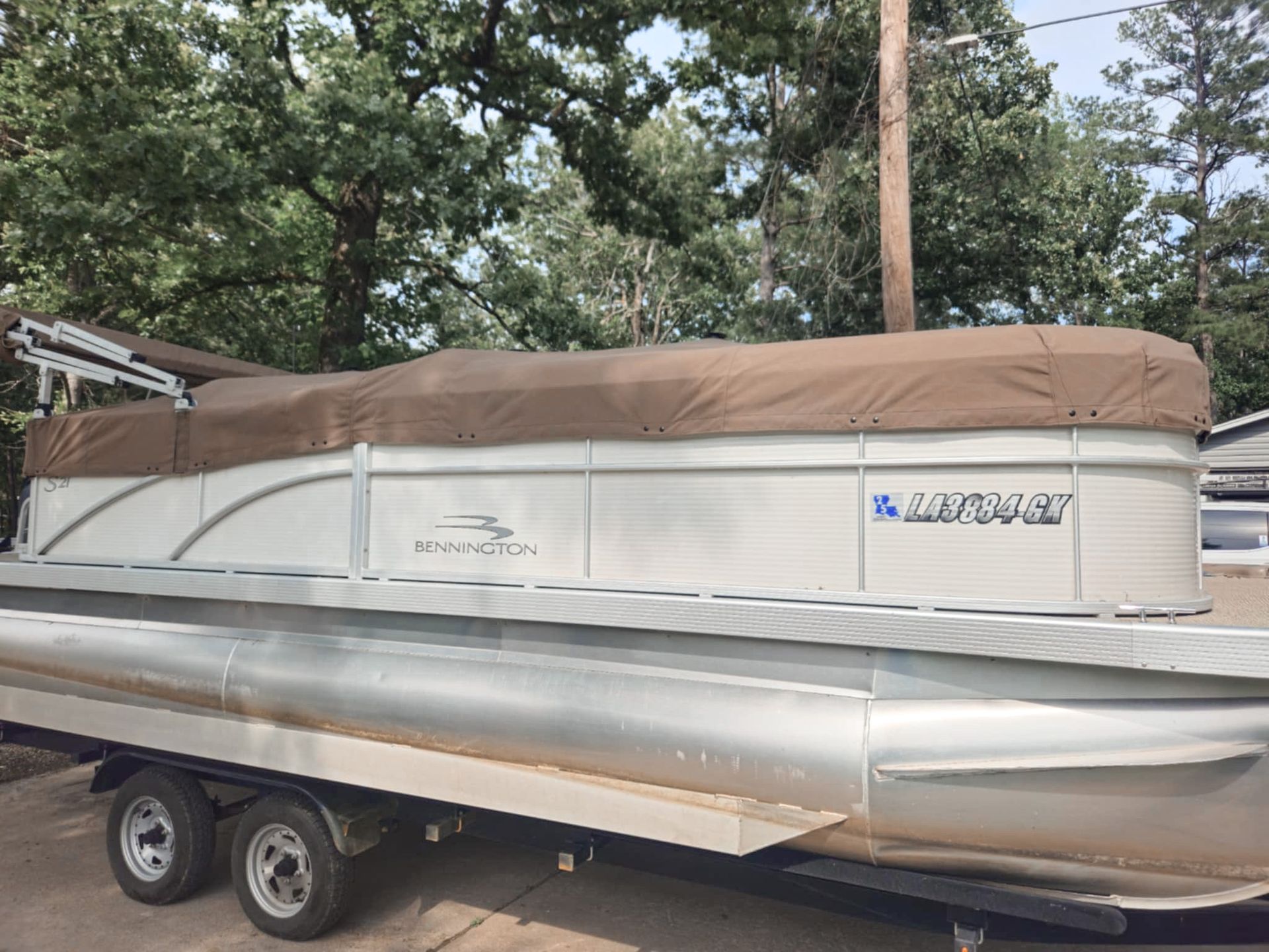 Pontoon boat on a trailer with a brown cover, parked outside near trees.