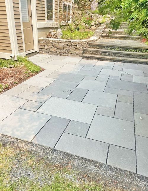 Gray stone patio beside a house, with steps and a low retaining wall leading to a garden path.