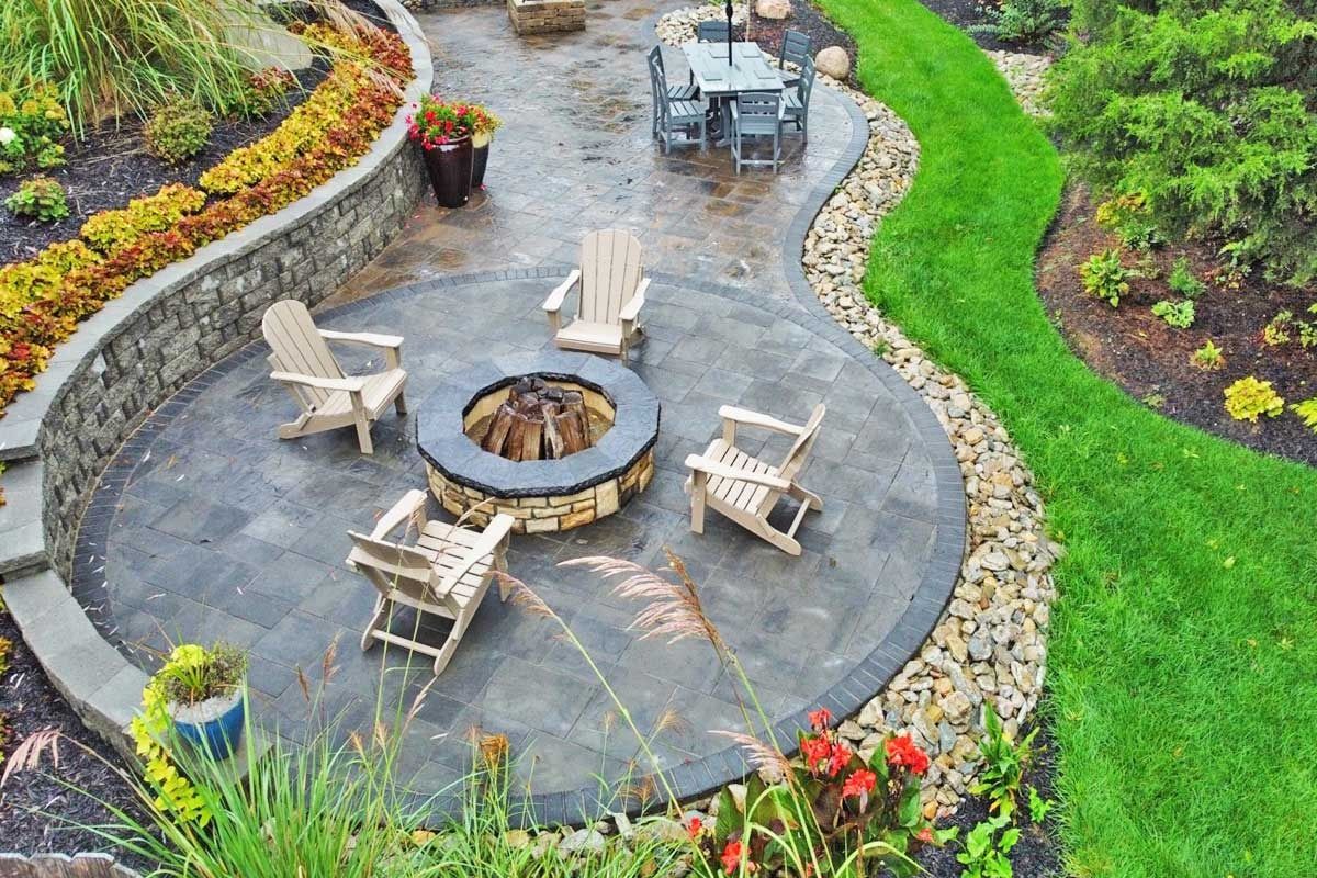 Stone patio with outdoor fire pit and wooden chairs in a landscaped garden