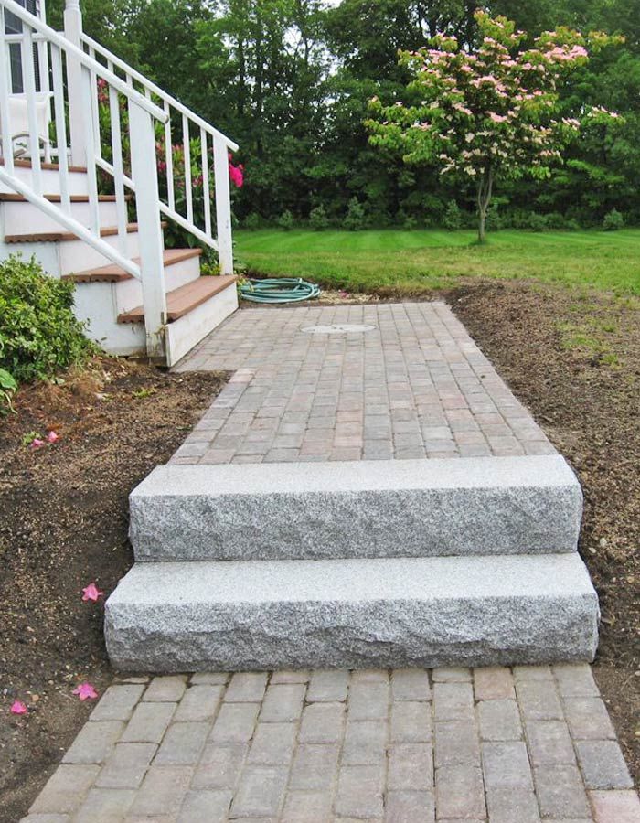 Brick walkway with two steps leading to a white porch, beside a green yard and bushes.