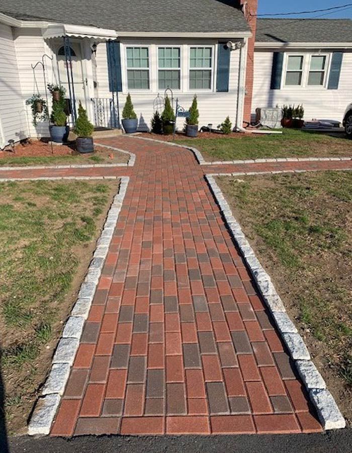 Brick walkway leading to a house entrance, bordered by concrete and flanked by small shrubs.