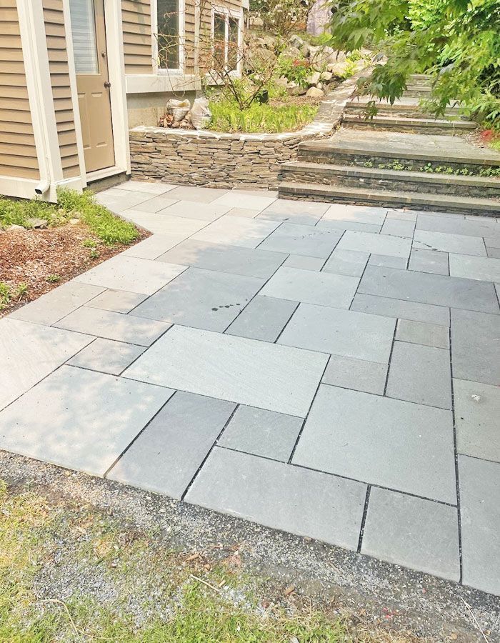 Gray paver patio beside a house, with stone steps and a low retaining wall in a landscaped yard