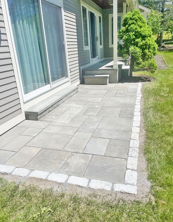 Patio of gray pavers beside a house with sliding glass doors and a grassy lawn edge