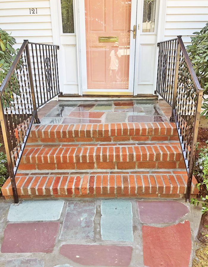 Brick front steps with iron railings leading to a white house door, with a multicolored stone walkway.