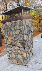 Stone chimney with a metal cap beside a paved walkway in an autumn wooded area