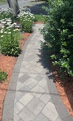 Curved gray tiled garden walkway bordered by white flowers, mulch, and green shrubs