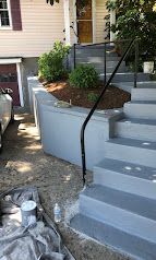 Outdoor concrete steps with black handrail beside a curved retaining wall and garden bed.
