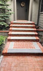 Brick front steps and walkway leading to a gray house door, bordered by red brick edging and greenery.