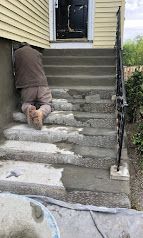 Person crawling up unfinished concrete stairs toward a house doorway with a handrail.
