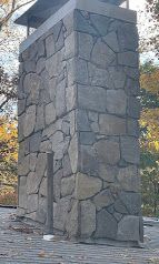 Stone monument or chimney with a rough wooden door, set on a paved surface outdoors among trees.