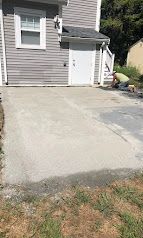Concrete driveway beside a gray house with white trim and a small shed door, bordered by grass.