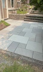 Paver patio with gray stone tiles beside a house, leading to stone steps and a landscaped garden area