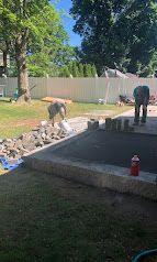 Two workers at a backyard pool construction site beside a gravel pit and fenced yard.
