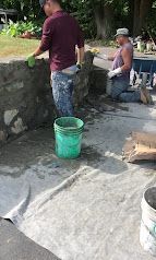 Two people working near a stone wall; one stands by a green bucket in a paved outdoor area.