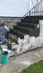 Worker painting a front staircase black beside a stone house wall