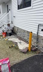 Worker installing stone veneer along the foundation of a white house beside a porch stairway.