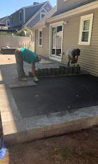 Two workers installing a black paver patio beside a beige house with sliding glass doors.
