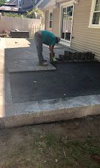 Worker smoothing a concrete patio beside a house with a dark asphalt area.