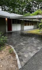 Modern house entrance with gray tiled driveway, red door, and large windows amid trees