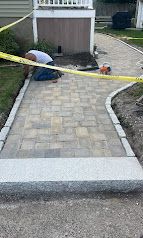 Worker laying pavers on a narrow walkway beside a house, with yellow caution tape across the path.