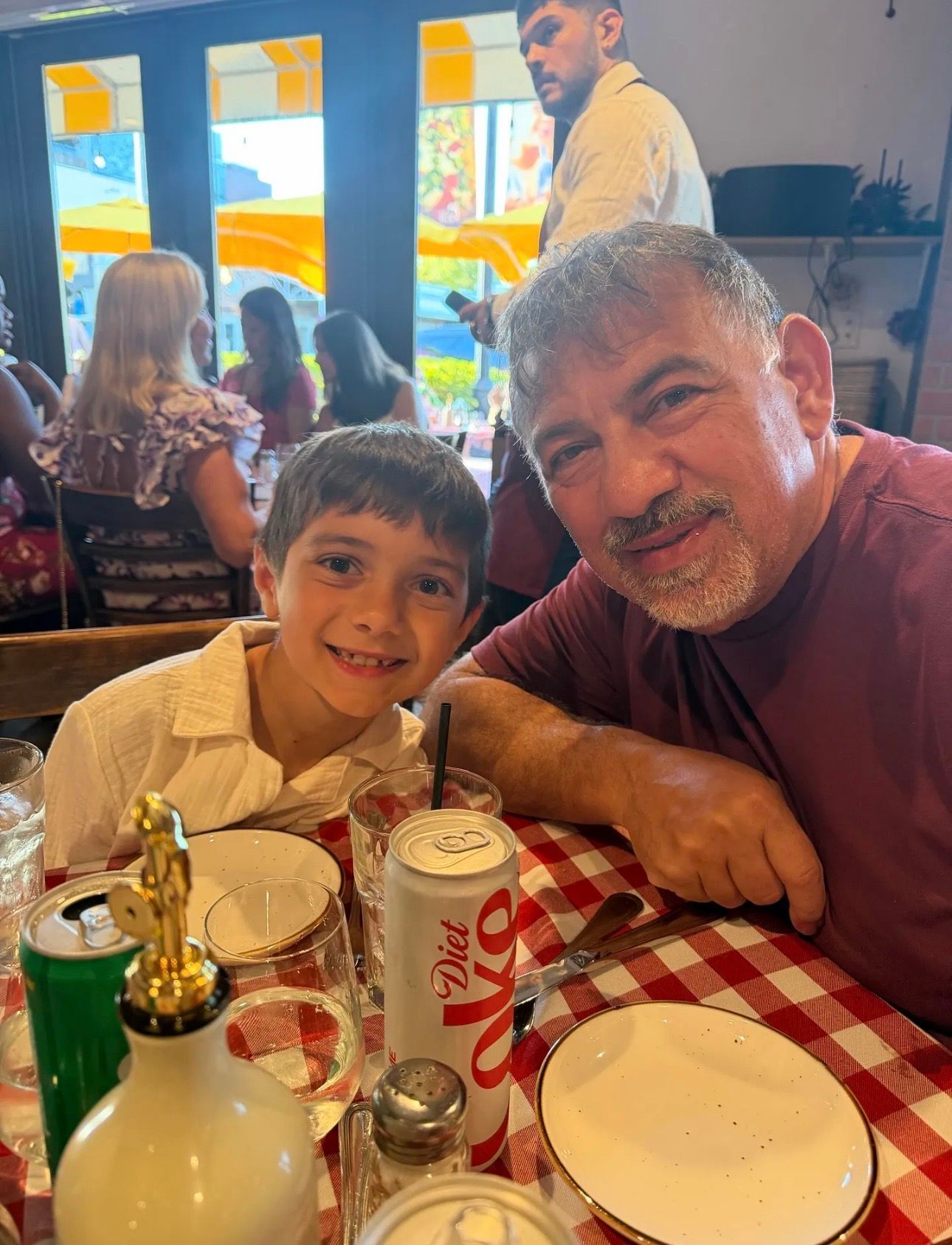 Two people smiling at a restaurant table with red-checkered cloth, desserts, and a soda can