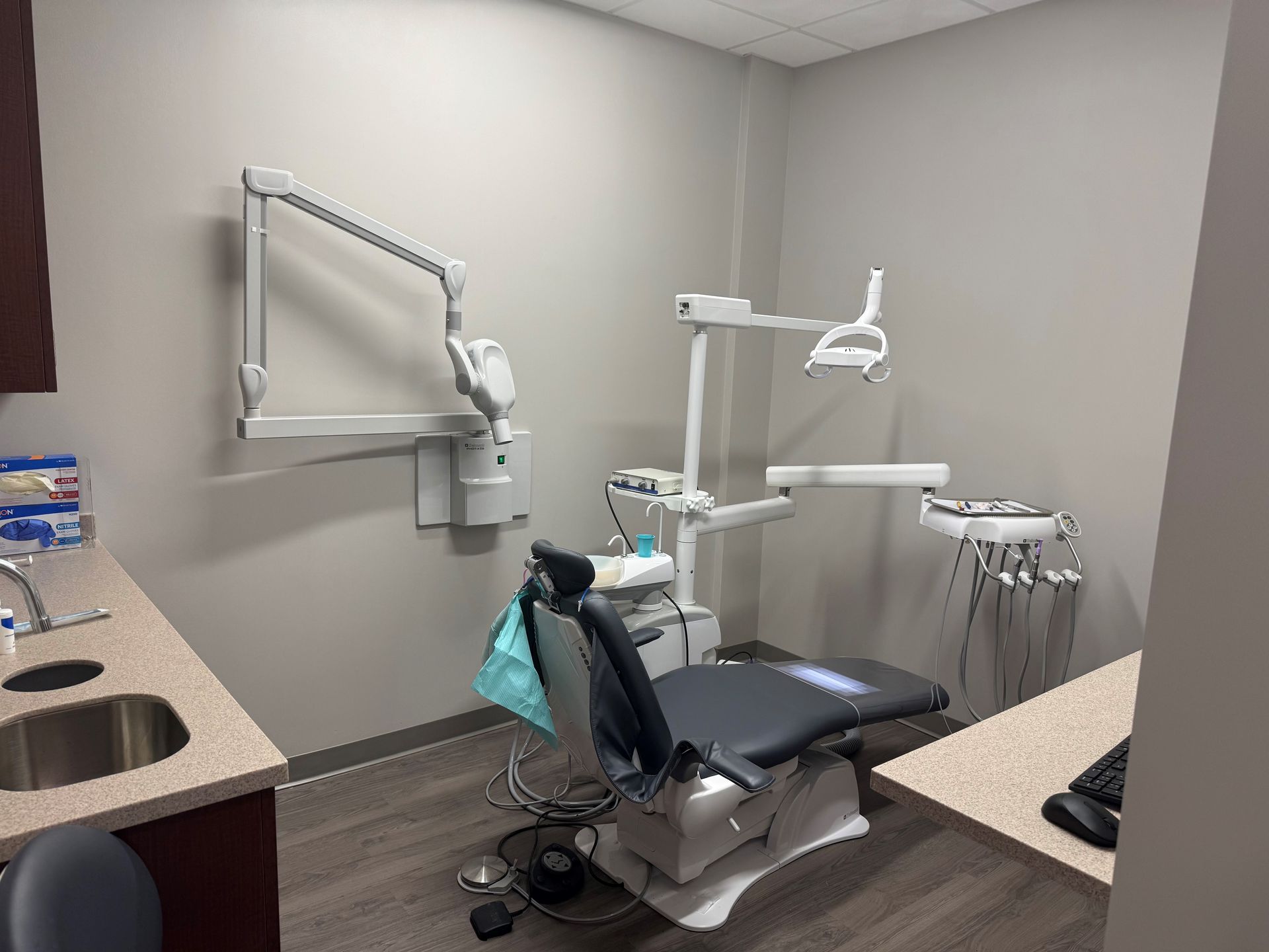 Dental exam room: dental chair, light, instruments, sink, gray walls, and desk.