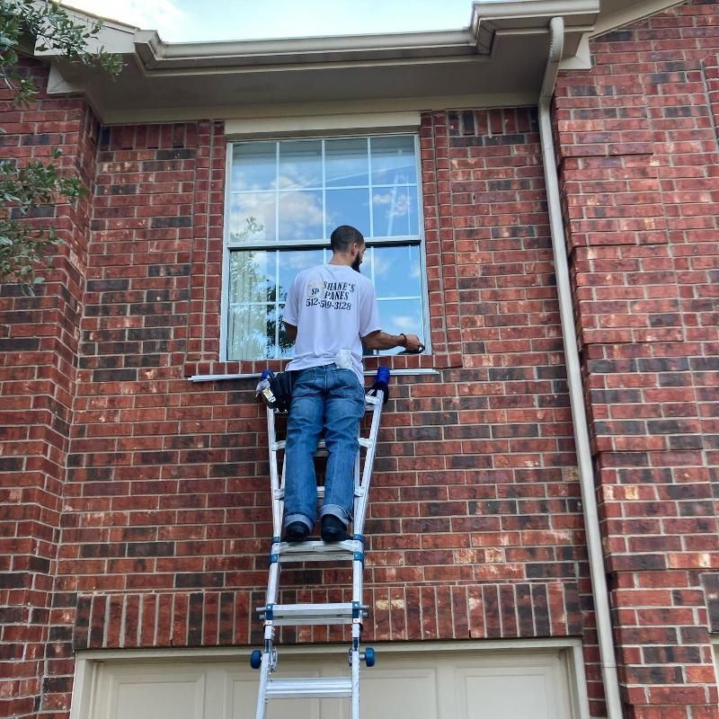 A man is standing on a ladder in front of a brick building cleaning a window.