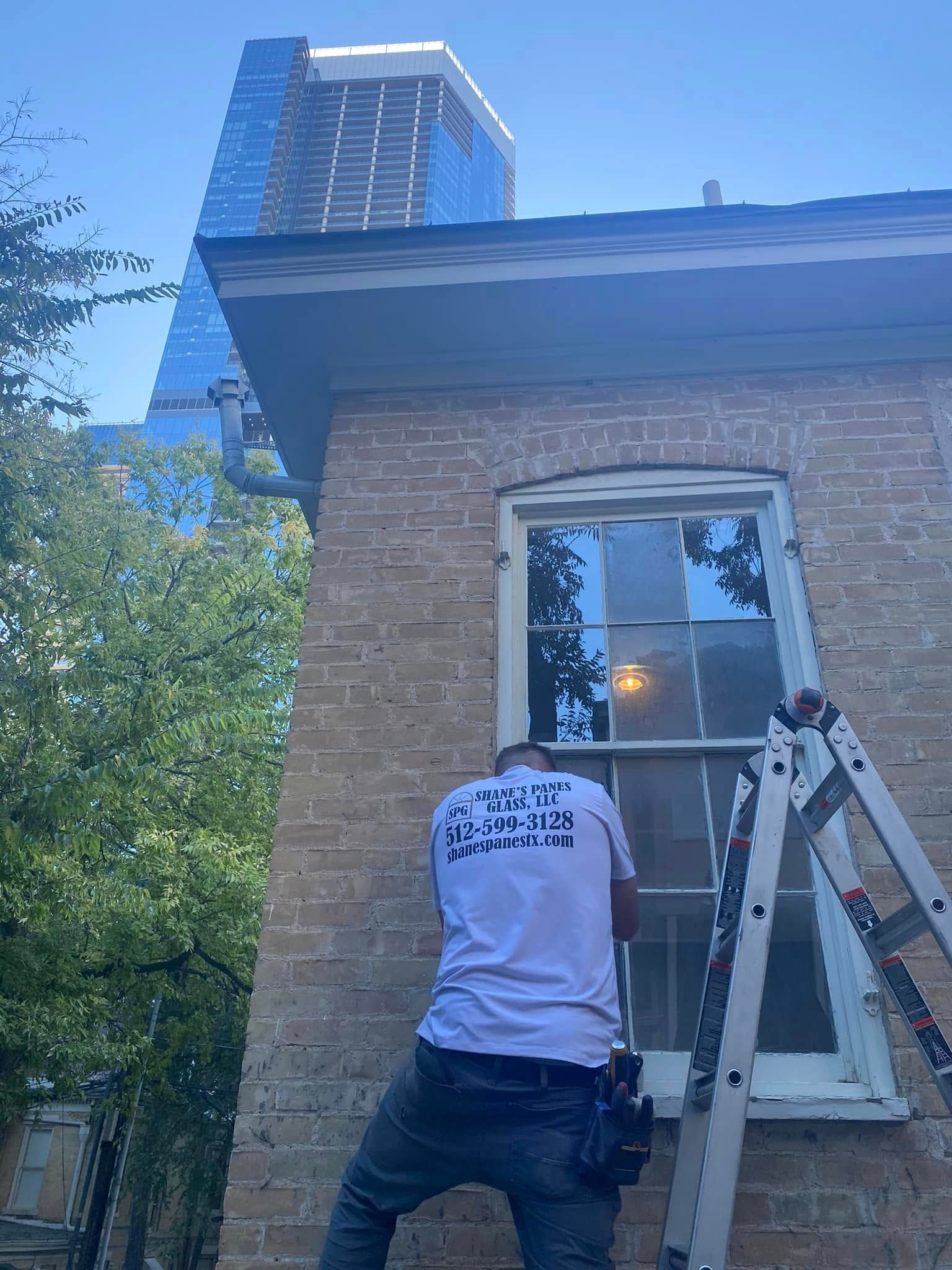 A man is standing on a ladder next to a window on a brick building.